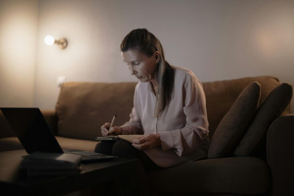 Older woman sitting on couch studying with pen, paper, and laptop for medical billing certification requirements. | medical billing certification requirements