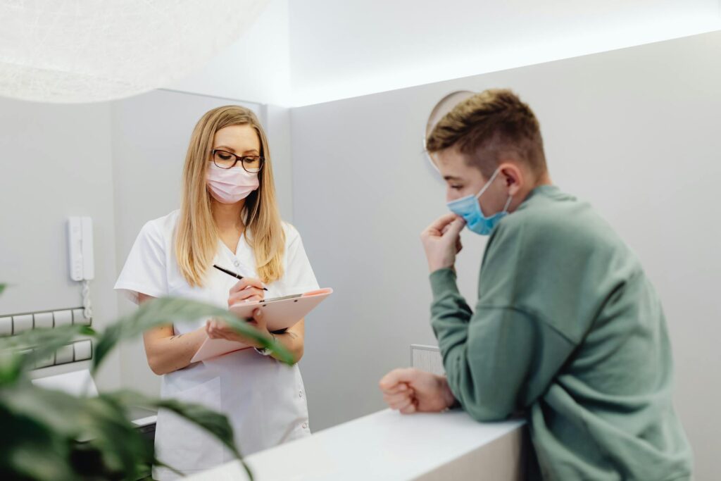 Healthcare professional in white coat and mask taking notes on a clipboard while consulting with a patient wearing a blue mask in a medical office setting, emphasizing communication and transparency in medical billing compliance.